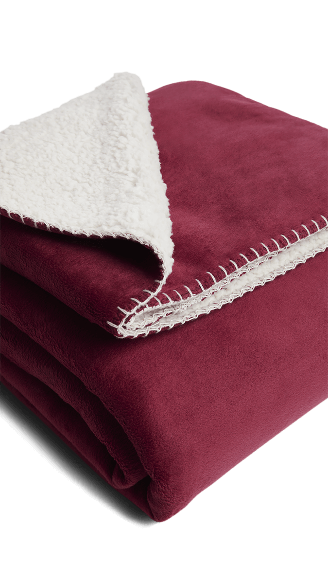 Gefaltete Sofadecke in Bordeauxrot mit weicher, weißer Sherpa-Fleece-Innenseite und dekorativer Ziernaht – ideal für gemütliche Stunden auf dem Sofa