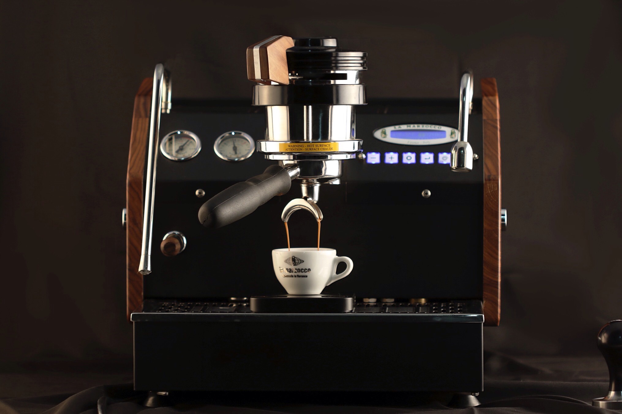 Eine La Marzocco Espressomaschine in edlem Schwarz mit Holzdetails zieht im stilvollen Setup alle Blicke auf sich. Die professionelle Siebträgermaschine brüht gerade einen doppelten Espresso direkt in die klassische La Marzocco Tasse. Ausgestattet mit Dualboiler, präziser Brühgruppe, digitalem Display und Barista-Steuerung – ideal für Kaffee-Enthusiasten, professionelle Baristas und anspruchsvolle Café-Umgebungen. Maximale Kontrolle für perfekte Kaffeezubereitung.