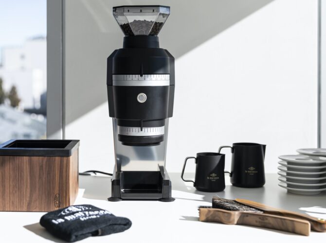 La Marzocco Swan Espressomühle in stilvoller Studio-Umgebung – professionelle Single-Dosing-Mühle mit präziser Mahlgradskala, mattschwarzem Gehäuse und hochwertiger Verarbeitung. Umgeben von passendem Barista-Zubehör wie Knockbox aus Holz, Milchkännchen, Tassen und Reinigungsbürsten – ideal für den Einsatz in Cafés oder Showrooms.