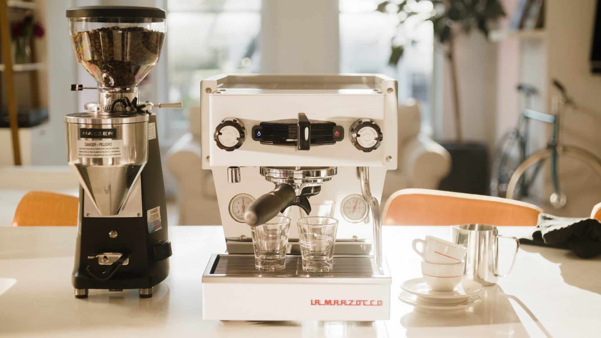 Weiße La Marzocco Siebträgermaschine mit Espressomühle auf heller Küchenzeile, zwei Espressogläser im Bezug, professionelle Espressomaschine für Gastronomie und anspruchsvollen Kaffeegenuss zu Hause